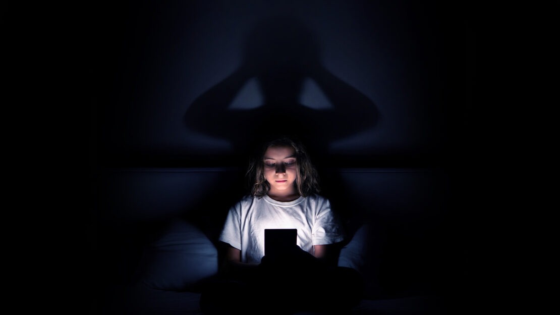 Minore al buio con smartphone e ombra inquietante: simbolo della dipendenza da social.