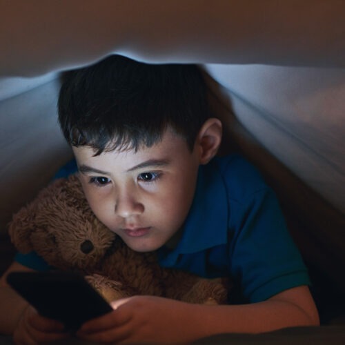 Bambino che usa smartphone sotto le coperte con un orsacchiotto, rappresentazione del rischio social per i minori under 14.