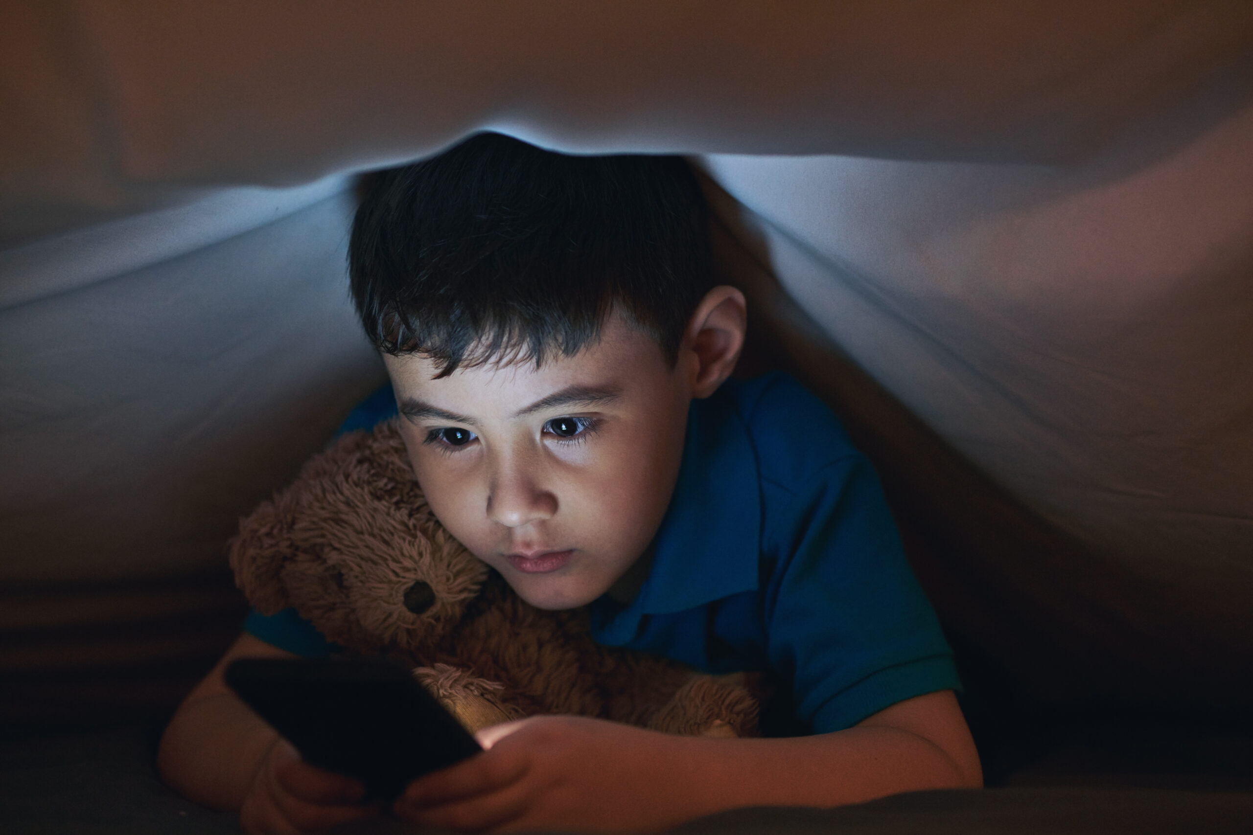 Bambino che usa smartphone sotto le coperte con un orsacchiotto, rappresentazione del rischio social per i minori under 14.