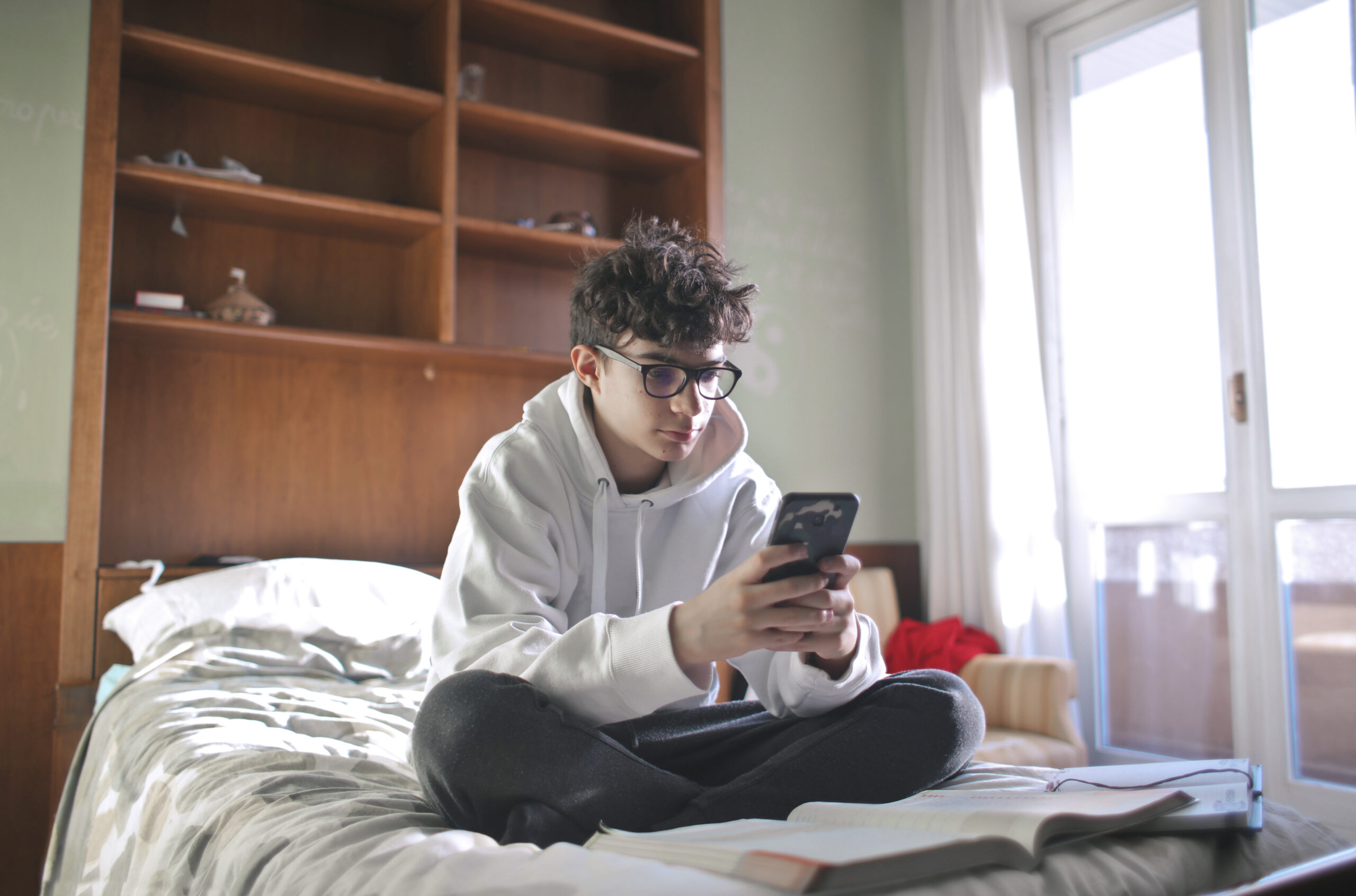 Ragazzo con occhiali e felpa seduto sul letto mentre usa lo smartphone con libri aperti: rappresentazione del cattivo uso dei social media e della gestione dei dati personali tra i minori in Italia. 