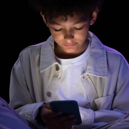 Ragazzo seduto da solo al buio mentre guarda lo smartphone, simbolo di isolamento sociale e dipendenza dai social nei minori
