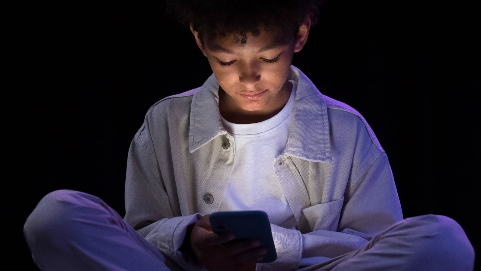 Ragazzo seduto da solo al buio mentre guarda lo smartphone, simbolo di isolamento sociale e dipendenza dai social nei minori